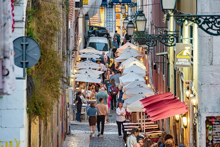 A narrow Bairro Alto street in Lisbon at dusk, crowded with people walking between rows of restaurant tables under white and red parasols, with lit street lanterns overhead