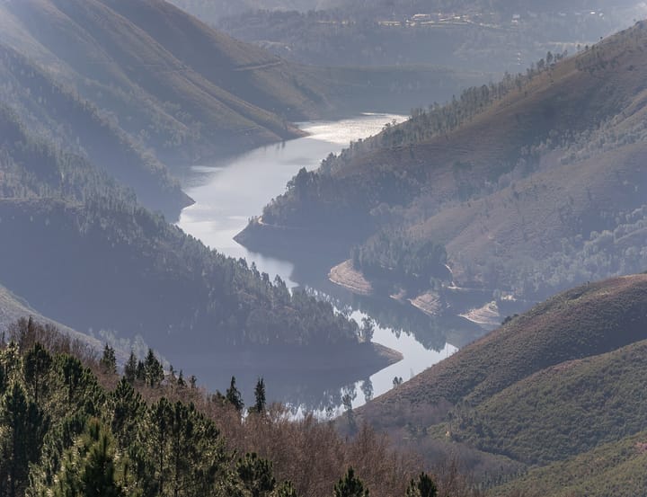 An aerial view of the Alto Lindoso reservoir in northern Portugal winding between forested hills, with pale drawdown lines marking the exposed shoreline above the water.