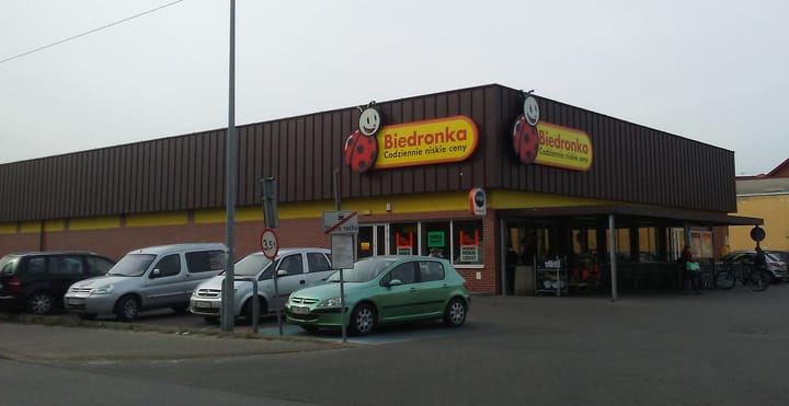 A Biedronka discount supermarket on a street corner in Poland, its yellow and red ladybird signs above a brick and brown-clad frontage, with cars parked outside