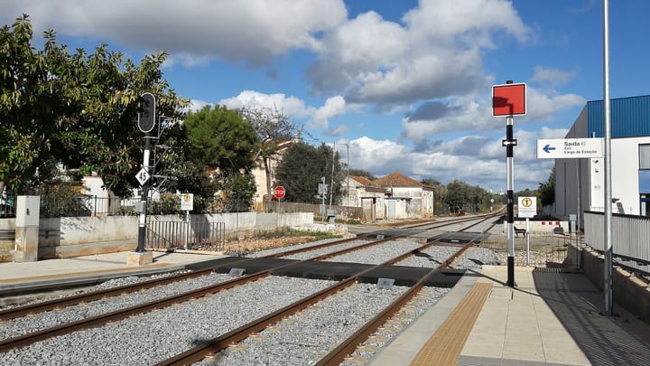 A level crossing beside Silves station in the Algarve, with rails running across a black rubber road surface, a red signal board and a blue Saida exit sign on the platform