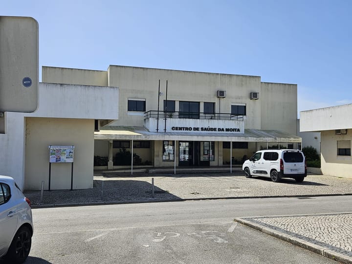 The low cream-coloured frontage of the Centro de Saude da Moita health centre in Portugal, its name in black letters above a canopy, with a van parked on the forecourt