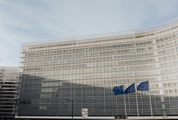 The curved glass and steel facade of the Berlaymont building in Brussels, headquarters of the European Commission, with three European Union flags flying in front of it under a pale sky.