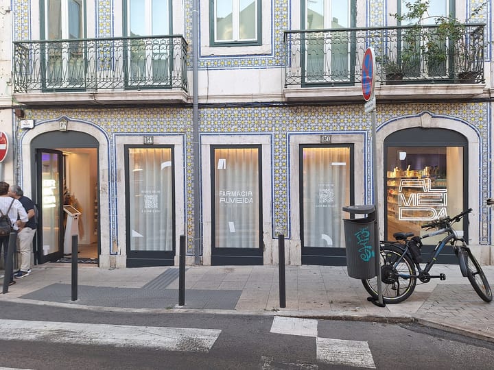 The tiled facade of Farmacia Almeida in Lisbon, its blue and yellow azulejos framing four arched shopfronts, with a bicycle parked at the kerb and customers at the open door.