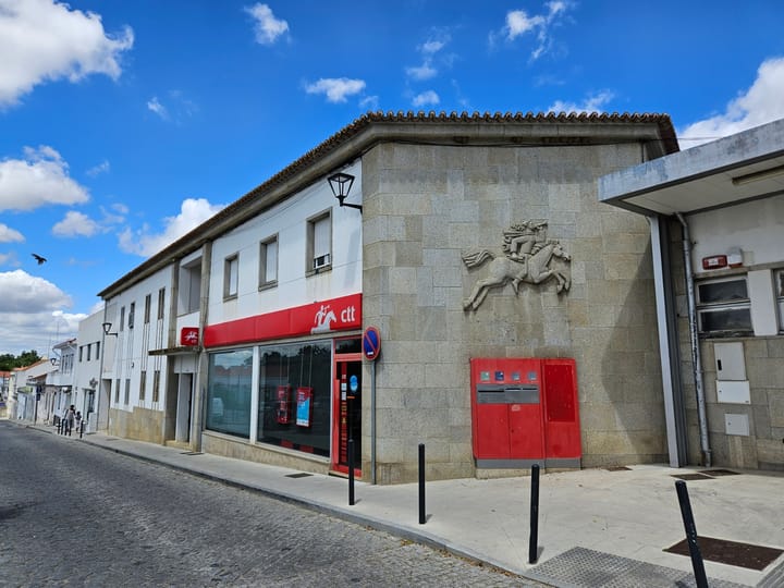 A CTT post office branch in Portugal with red signage and a red post box set into a granite wall below a stone relief of a mounted postilion
