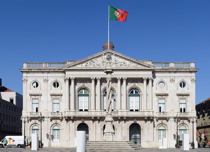 The neoclassical facade of the Pacos do Concelho, Lisbon's city hall, with the Portuguese national flag flying from the flagpole above its dome against a clear blue sky.