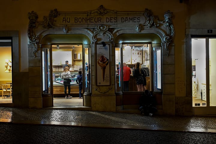An ornate carved stone shopfront in central Lisbon at night, its lintel reading Au Bonheur des Dames, the two arched windows now lit as an ice cream counter with customers inside.