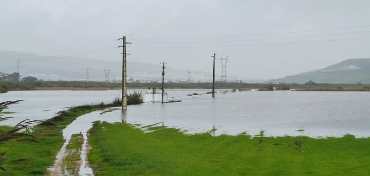 Floodwater covering a Portuguese river valley up to the base of electricity pylons and poles, with a town and green hills visible through the rain haze beyond