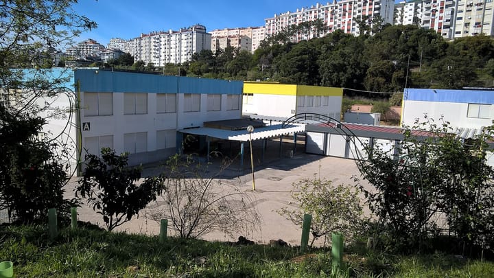 A Portuguese state basic school of low white blocks with blue and yellow trim and a covered walkway, with tall residential apartment towers on the hillside behind