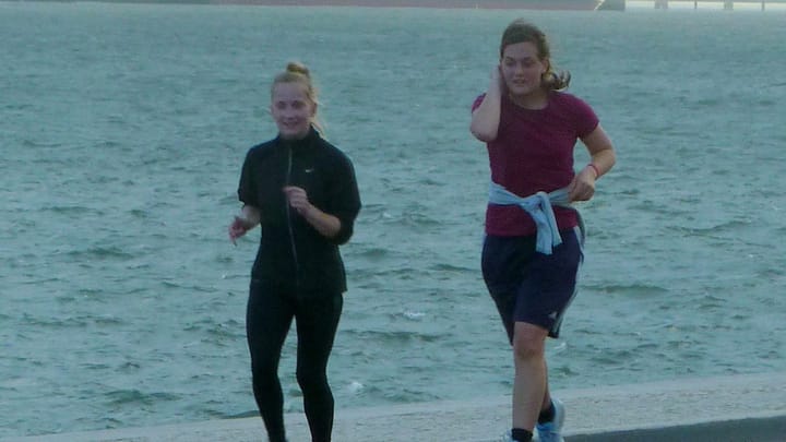 Two runners on the paved riverside path at Belem in Lisbon, with the grey water of the Tagus estuary behind them