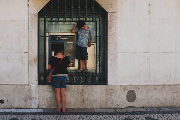 A woman and a child at a wall-mounted Multibanco cash machine set in a stone facade on a Lisbon street, the child standing on the ledge to reach the screen.