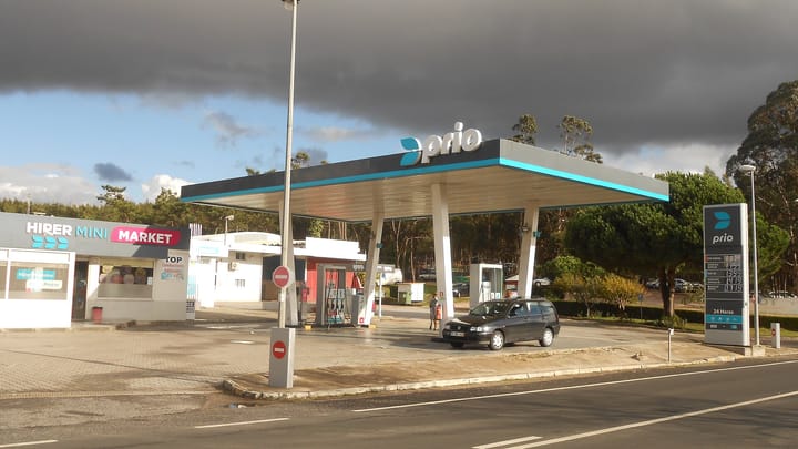 A Prio filling station on a Portuguese roadside with a car at the pumps under the canopy and a price board listing diesel and petrol prices in euros
