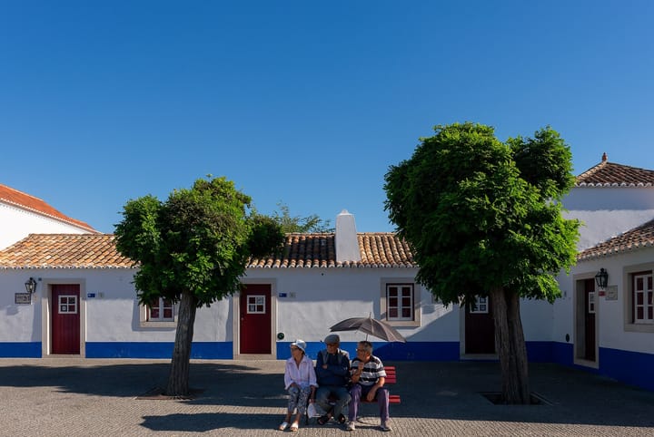 Three older people sit together on a red bench in the square at Porto Covo, Portugal, in front of whitewashed single-storey houses with blue skirting and red doors.