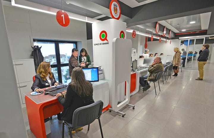 The interior of a Portuguese Espaço do Cidadão public service office, with staff at numbered red and white desks serving citizens and others waiting on chairs along the wall.
