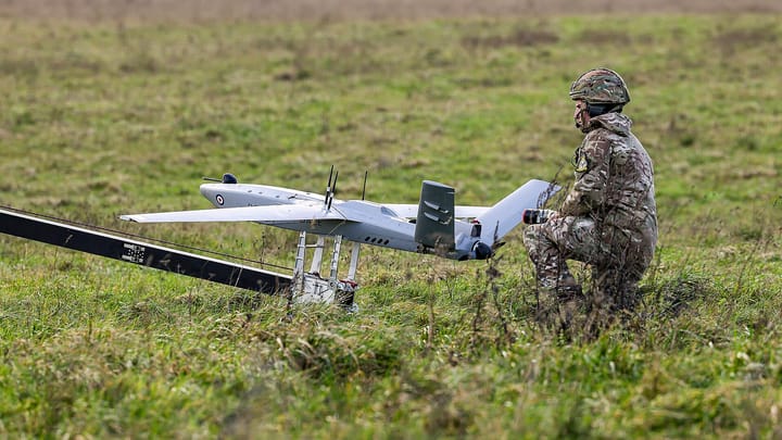 A Tekever AR3 fixed-wing drone in Royal Air Force markings sits on a catapult launch rail in a grass field, with a serviceman in camouflage crouched beside it.