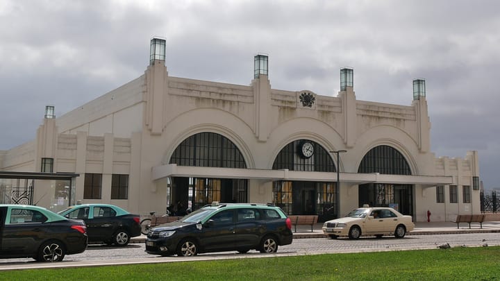 The white art deco facade of the Terreiro do Paço ferry terminal in Lisbon, with three tall arched windows, a clock above the central arch and Lisbon taxis parked along the kerb in front.