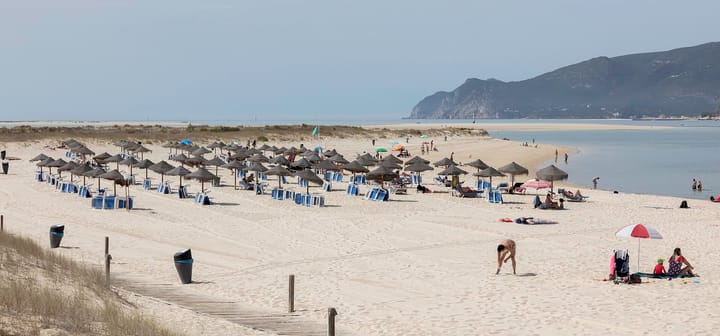 Rows of straw parasols and blue sunbeds on the wide sand of Troia beach in Portugal, with a wooden boardwalk in the foreground and the Arrabida hills across the water behind.