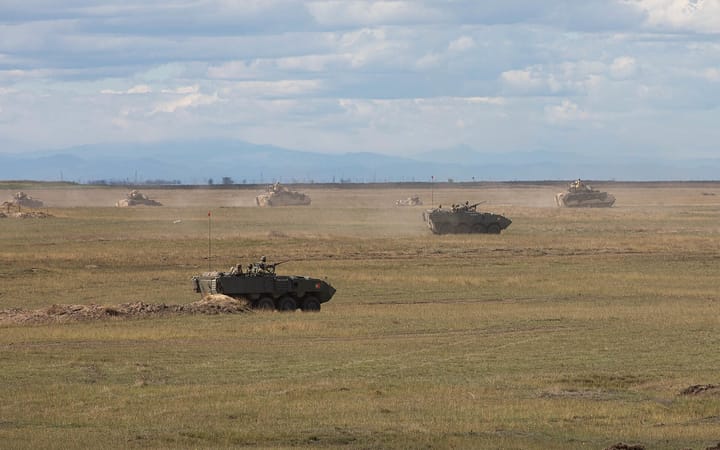 A Portuguese Army Pandur II wheeled armoured vehicle with the national flag on its hull, halted on open ground during a NATO exercise as other vehicles raise dust behind it