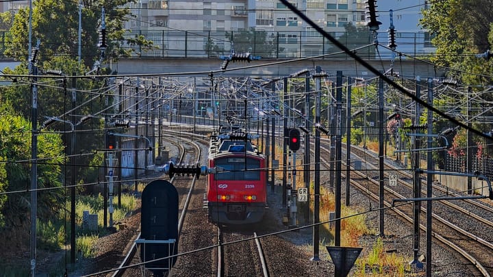 A red CP suburban train on the Sintra line approaching through a web of overhead wires at Benfica, Lisbon, with apartment blocks behind