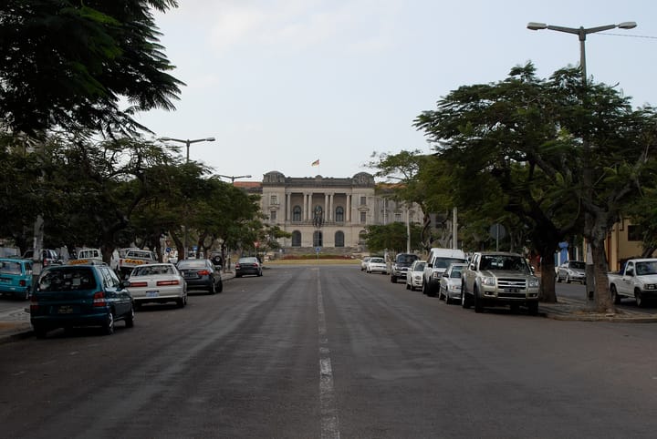 Avenida Samora Machel in Maputo looking towards the colonnaded city hall, with the Mozambican flag flying above it and cars parked under the trees on both sides