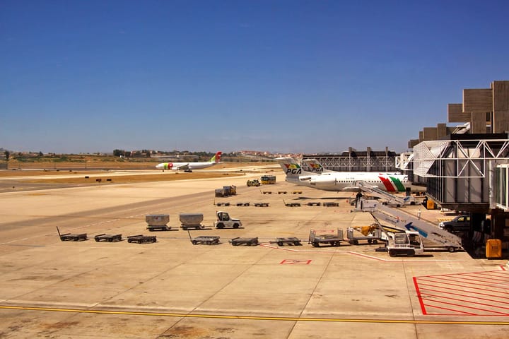 Baggage dollies, a Groundforce tug and passenger steps lined up on the apron at Lisbon Airport beside a jet bridge, with a TAP aircraft taxiing in the background