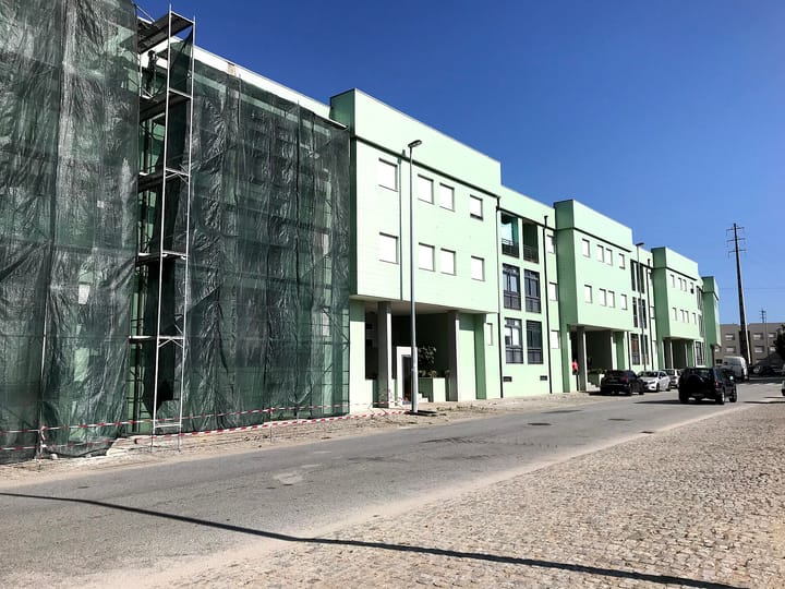 A row of pale green three-storey apartment blocks on a street in Rio Tinto near Porto, with one end wrapped in scaffolding and green netting and cars parked along the kerb
