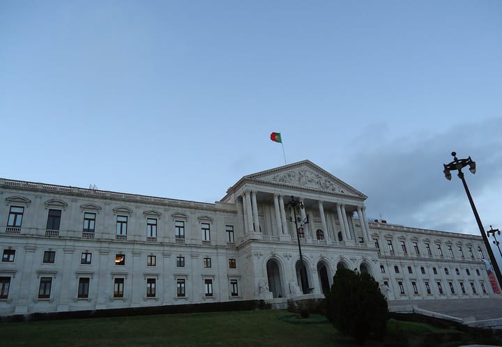 The neoclassical white facade of the Palacio de Sao Bento in Lisbon at dusk, with the Portuguese flag flying above the columned pediment and a lamp post in the foreground
