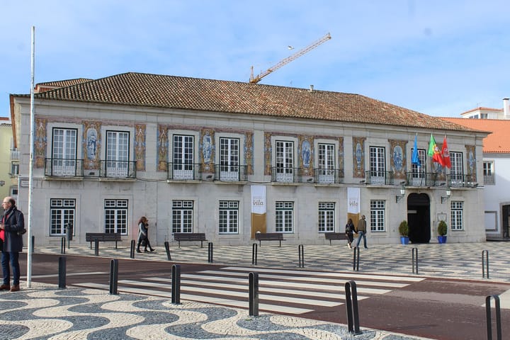 The azulejo-panelled front of the Cascais town hall on a cobbled square, with Portuguese and European flags at the door and calcada paving in the foreground