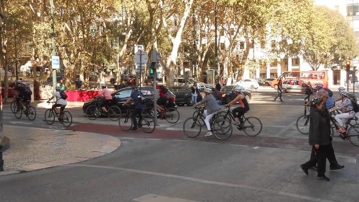 Cyclists crossing a red-surfaced cycle lane on Lisbon's Avenida da Liberdade under plane trees, with pedestrians walking over calcada paving in the foreground