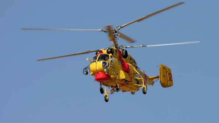 A yellow Kamov Ka-32 firefighting helicopter of the Portuguese fire brigade in flight against a clear blue sky, with red water tanks along its fuselage