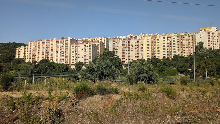 A row of tall cream and pink apartment blocks on a wooded hillside on the outskirts of Lisbon, seen across scrubland beside a road