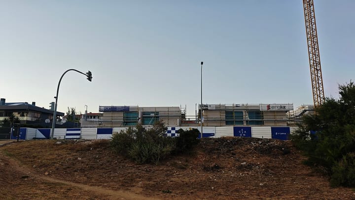 A low-rise residential development under construction in Sao Pedro do Estoril, with scaffolding, hoarding along the site and a yellow tower crane against an evening sky