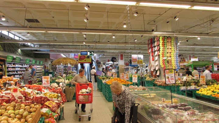 Shoppers with red trolleys and baskets in the fruit and vegetable section of a Continente supermarket in Albufeira, with crates of oranges, lemons and melons
