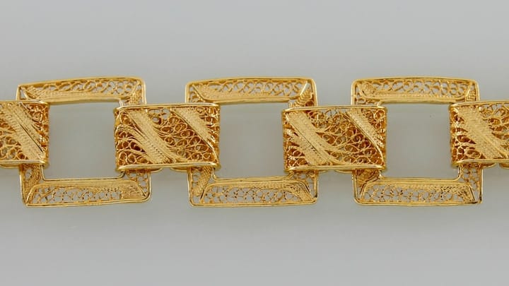 Close-up of a Portuguese gold filigree bracelet made of linked square frames filled with fine twisted wire scrolls, on a pale grey background