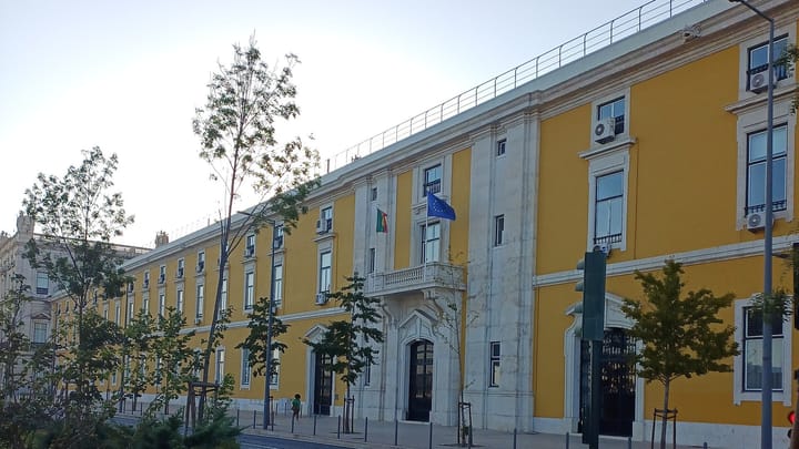 The long yellow neoclassical facade of the Ministry of Finance headquarters in Lisbon, with Portuguese and European Union flags flying from a first-floor balcony