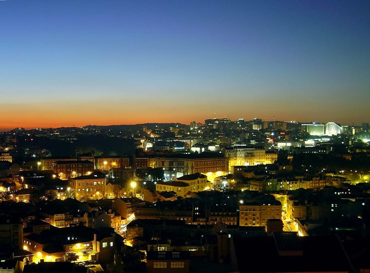 A panorama of Lisbon at dusk from the Miradouro da Senhora do Monte, with lit streets below and an orange band of light along the horizon