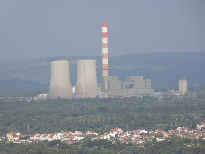 The Pego power station in central Portugal seen from a distance, its two cooling towers and red and white striped chimney rising behind wooded hills and a village of white houses