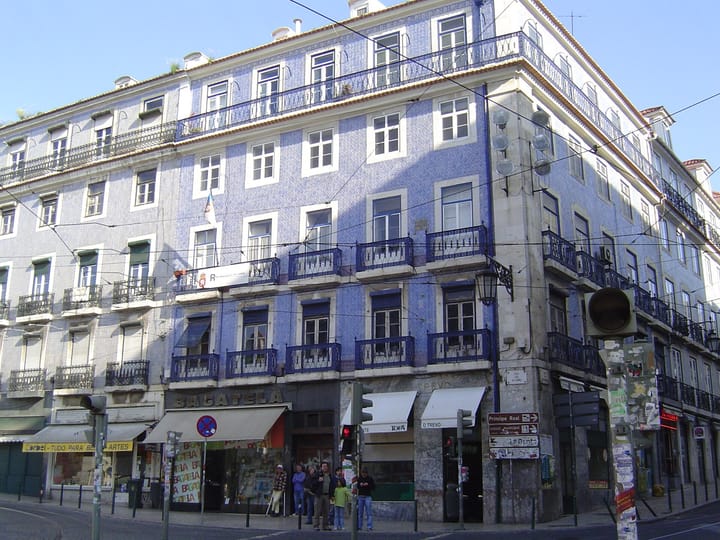 A corner apartment building in Lisbon faced in blue azulejo tiles, with wrought-iron balconies on every floor and small shops at street level