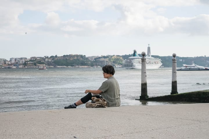 A visitor sitting on the steps of the Cais das Colunas in Lisbon looking out at the Tagus, with a cruise ship on the water and the far bank at Almada behind