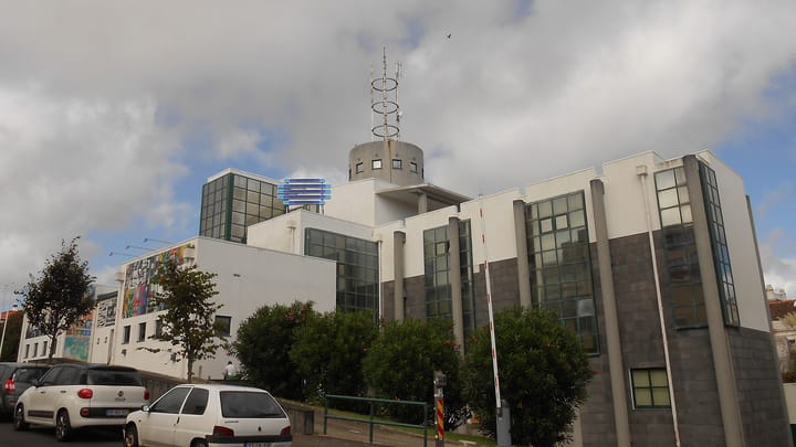 The RTP regional broadcasting centre in Ponta Delgada in the Azores, a white and grey building with the blue RTP logo on its wall and a transmission mast above the roof