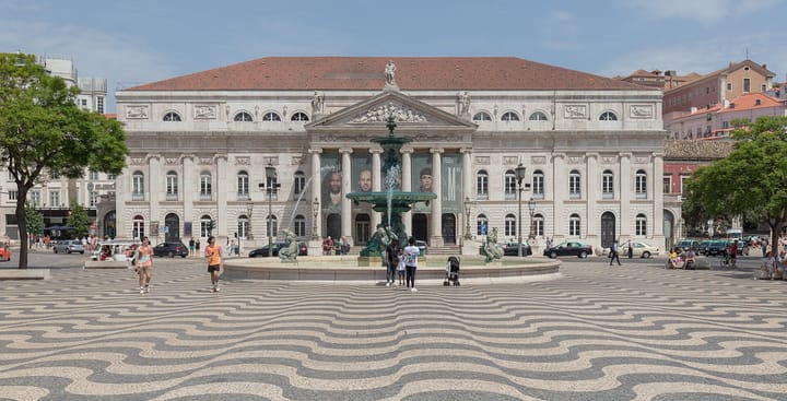 The Teatro Nacional D. Maria II on Lisbon's Rossio square, its columned neoclassical facade behind the bronze fountain and the wave-patterned calcada paving