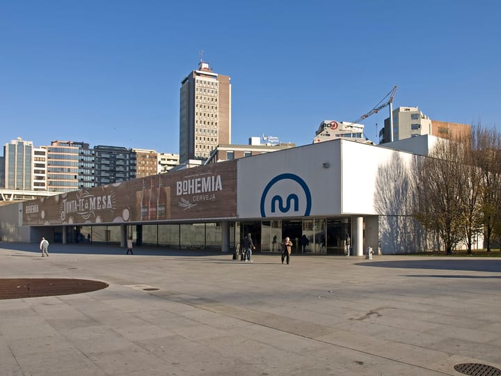 The exterior of the Trindade metro station in Porto, its low white concourse carrying the blue Metro do Porto logo, seen across the paved forecourt