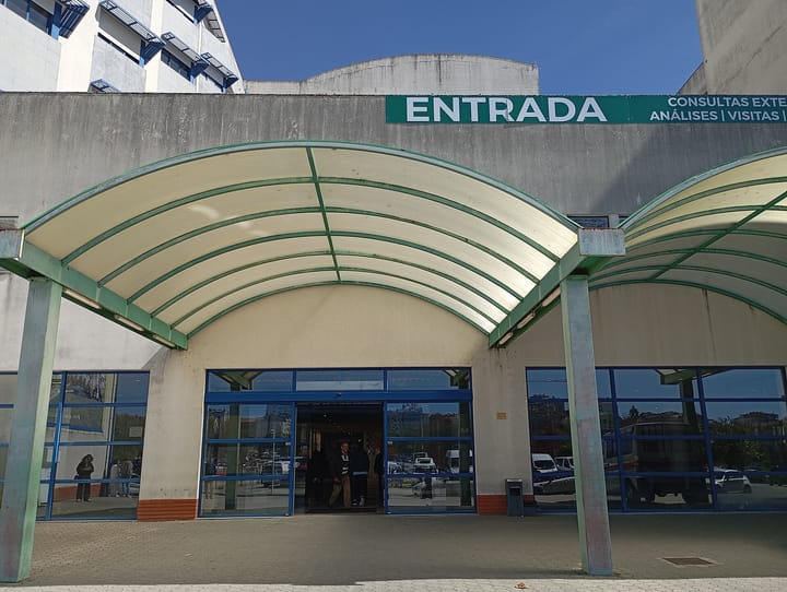 The main entrance of a Portuguese public hospital under a curved canopy, with a green sign reading Entrada and Consultas Externas, Análises and Visitas