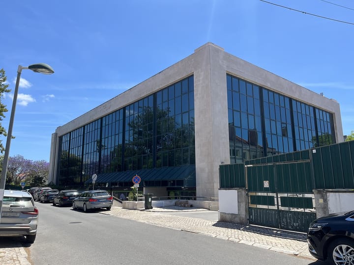 A modern stone and glass office building on a quiet street in Belém, Lisbon, with parked cars along the kerb and a jacaranda in bloom to one side