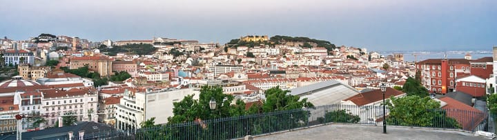 A wide view over central Lisbon at dusk from the Miradouro da Senhora do Monte, red rooftops running down to the Tagus with the Castelo de Sao Jorge on the skyline
