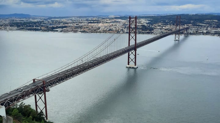 The Ponte 25 de Abril seen from the Almada side, its red steel towers and suspended roadway crossing the Tagus towards Lisbon under a cloudy sky