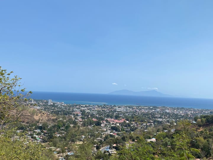 Dili seen from a hillside above the city, its low roofs spread along the coast with the blue sea and a distant island on the horizon