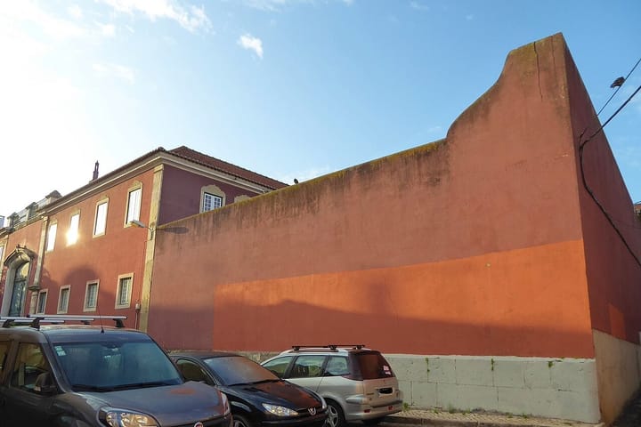 Old terracotta-red buildings and a high rendered wall along the Rua de Campo de Ourique in Lisbon, with cars parked along the kerb in late afternoon sun