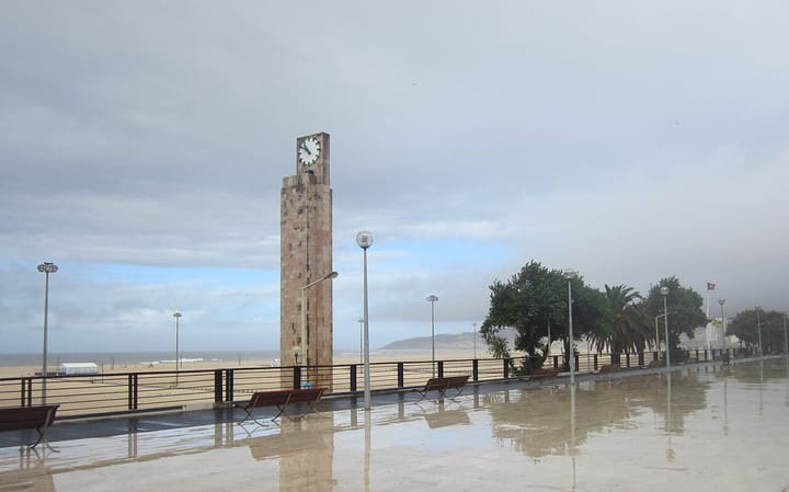 The clock tower on the seafront esplanade at Figueira da Foz, with benches, a wet promenade and the wide sandy beach under a grey sky