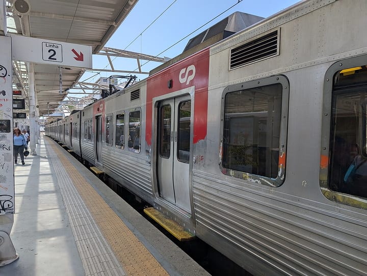 A silver CP suburban train with red door panels standing at platform 2 of Roma-Areeiro station in Lisbon under a clear blue sky