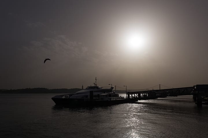 A ferry moored at the Cais do Sodré pier in Lisbon in hazy evening light, with a gull overhead and the Ponte 25 de Abril faint in the distance